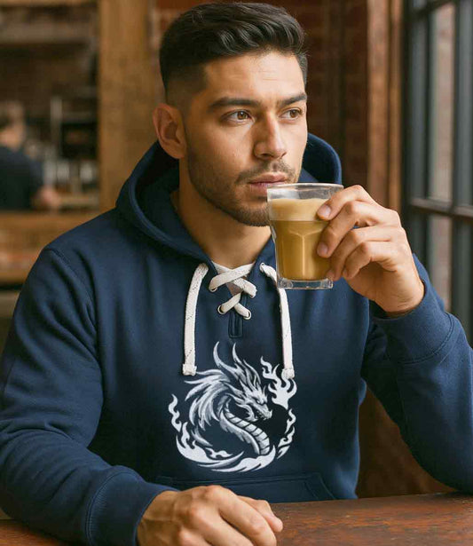 Man in a blue hoodie with a dragon design drinking coffee in a cozy setting.